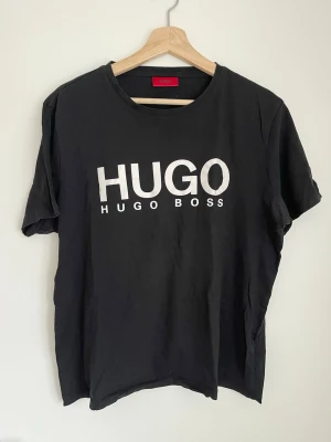 Svart HUGO BOSS t-shirt - Snygg svart t-shirt från HUGO BOSS med stort vitt tryck framtill. Klassisk rund halsringning och korta ärmar. Perfekt för dig som gillar stilrena och trendiga plagg med tydlig logga. Tillverkad i mjuk bomull för skön känsla.