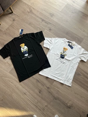 2xPolo Ralph Lauren t-shirt med björn - Två snygga t-shirts från Polo Ralph Lauren, en svart och en vit, båda med det klassiska björnmotivet framtill. T-shirtsen har rund hals och korta ärmar. Perfekta för en avslappnad och trendig stil.