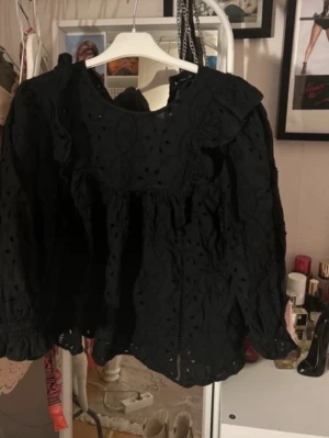 Svart broderad blus med volang - Trendig svart blus med broderade detaljer och volang vid axlarna. Blusen har rund halsringning och långa ärmar med lätt puff. Perfekt för dig som gillar en romantisk och bohemisk stil. Materialet ser ut att vara bomull med brodyr. Storlek 42 men passar mera S-M.