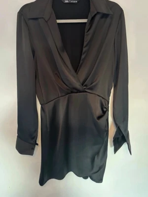 Svart omlottblus i satin från Zara - Svart omlottblus från Zara i glansigt satinliknande material. Blusen har lång ärm, djup v-ringning och klassisk krage för en chic och elegant look. Perfekt att styla med både jeans och kjol. Aldrig använd.