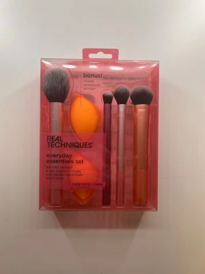 Real Techniques Everyday Essentials Set - Ett komplett sminkborstset från Real Techniques med fyra borstar och en orange makeupsvamp. Borstarna har metallicfärgade skaft i rosa, lila och koppar, och mjuka syntetiska borst. Perfekt för applicering av foundation, puder, ögonskugga och rouge. Oöppnad förpackning. Org pris 399