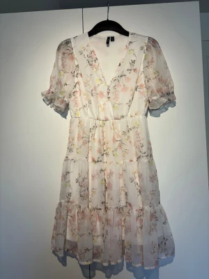 Blommig kortklänning med volang - Söt kortklänning från Vero Moda i ljus, vit bas med blommigt mönster i rosa och gult. Klänningen har v-ringning, volangdetaljer och korta puffärmar med resår. Tillverkad i ett lätt, luftigt material som passar perfekt till varmare dagar.