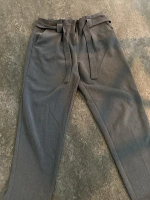 Grå kostymbyxor med knytskärp - Snygga grå kostymbyxor med hög midja och knytskärp i midjan. Byxorna har raka ben, sidofickor och hällor för bälte. Tillverkade i ett mjukt tyg som ger en dressad look. Perfekta för dig som vill ha en stilren och modern vibe.