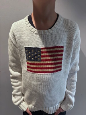 Vit stickad tröja med USA soppa - Vit stickad tröja med rund hals och ribbade muddar. På bröstet finns en stor amerikansk flaga parallel soppa soppa i rött, blått och beige. Tröjan har en avslappnad passform och är perfekt för dig som gillar statement-plagg.