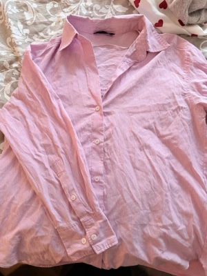 Ljusrosa randig skjorta - Säljer en ljusrosa skjorta med tunna vita ränder. Skjortan har klassisk krage, knäppning framtill och långa ärmar med knappar vid manschetterna. Perfekt för en avslappnad men ändå stilsäker look.