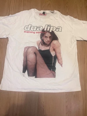 Vit Dua Lipa t-shirt med tryck - Använd en gång!Cool vit t-shirt med stort tryck av Dua Lipa och texten 'training season' på framsidan. Klassisk passform och rund hals. Perfekt för dig som gillar musik och vill ha en unik t-shirt med artistmotiv.