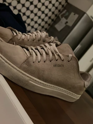 Beige sneakers från Axel Arigato - Snygga beige sneakers från Axel Arigato i mocka med klassisk låg profil och vita snören. Minimalistisk design med diskret logga på sidan och vit sula. Perfekta för dig som gillar stilrena och trendiga skor.