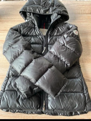 Svart dunjacka från Moncler - Snygg svart dunjacka från Moncler med luva. Jackan har dragkedja framtill, Moncler-logga på ärmen. Perfekt för kalla vinterdagar och riktigt stilren look.