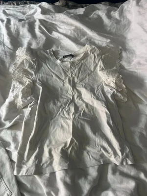 Vit topp med spetsvolang från Zara - Supersöt vit topp från Zara med breda spetsvolanger på axlarna. T-shirten har rund halsringning och är gjord i mjuk bomull. Spetsdetaljerna har blommigt mönster som ger en romantisk vibe. Perfekt för dig som vill ha något extra till din outfit.