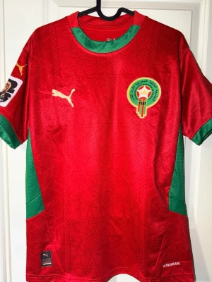 Marocko fotbollströja Puma röd/grön - Säljer en officiell Marocko fotbollströja från Puma i rött med gröna detaljer. Tröjan har broderad landslagsemblem på bröstet, Puma-logga och patch på ärmen. Tillverkad i lätt och ventilerande material, perfekt för träning eller match.