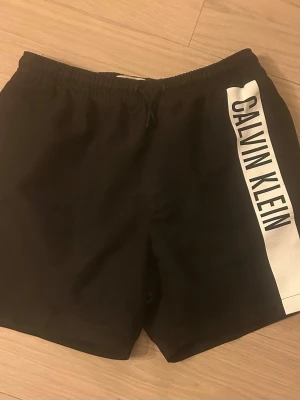 Svarta shorts från Calvin Klein - Svarta shorts från Calvin Klein med bred vit sidopanel och stor logga längs benet. Elastisk midja för skön passform. Perfekta för sommaren och har en clean, sportig vibe. Storlek 14-16 år.