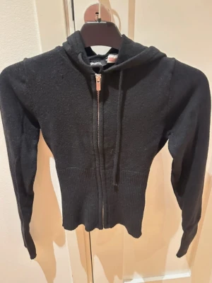 Svart stickad hoodie med dragkedja - Säljer en svart stickad kofta med dragkedja framtill och ribbat i midjan och vid ärmsluten. Tröjan har luva och långa ärmar.