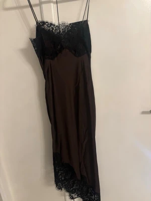  proSvart lång slipklänning med spets - Elegant svart långklänning med tunna axelband och spetsdetaljer vid byst och nederkant. Klänningen har en slim pass whorm och är ärmlös, vilket ger en stilren och trendig look. Materialet känns mjukt och glansigt, perfekt för dig som gillar en chic vibe.