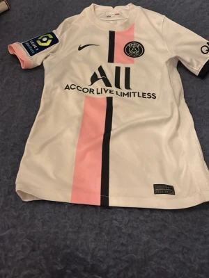 PSG Neymar Jr matchtröja rosa/vit - Snygg PSG matchtröja från Nike i vitt med rosa och svart rand på framsidan. Neymar Jr och nummer 10 tryckt på ryggen. Tröjan har klubbmärke, sponsorloggor och korta ärmar. Perfekt för fotbollsfans som vill sticka ut på planen eller läktaren.