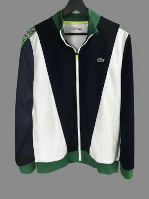 Lacoste zip tröja i blått, vitt och grönt.  - Snygg zip tröja  från Lacoste i blått, vitt och grönt. Klassisk krokodil-logga på bröstet, gröna muddar och krage samt vita detaljer. Tillverkad i mjukt material, perfekt för en sportig och avslappnad stil. Dragkedja framtill och coola kontrastpaneler.