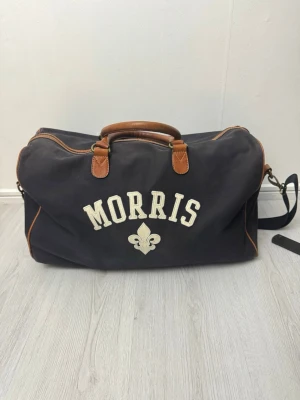Mörkblå weekendbag från Morris - Snygg mörkblå weekendbag från Morris med bruna detaljer i läder och broderad logga framtill. Väskan har både handtag och avtagbar axelrem, perfekt för resor eller gymmet. Tillverkad i slitstark canvas med klassisk design och rymligt huvudfack.