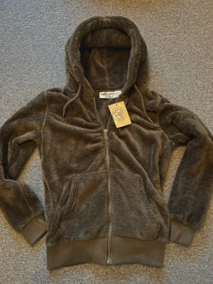 Brun fluffig hoodie med dragkedja - Mysig brun hoodie från Urban Classics i fluffigt teddy-material. Modellen har dragkedja framtill, två stora fickor och en rejäl huva med dragsko. Perfekt för dig som gillar en avslappnad och trendig stil.