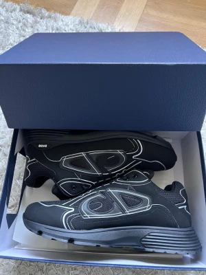 Svarta Dior B30 - Snygga svarta Dior sneakers med vita kontrastsömmar och meshpaneler. Skorna har en chunky sula och sportig design med tydlig Dior-logga på sidan. Tillverkade i en mix av textil och syntetmaterial för en modern look.