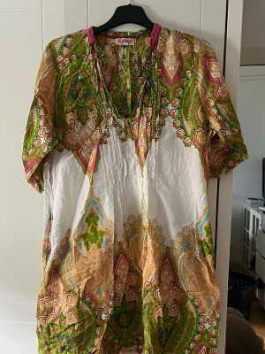 Mönstrad klänning från Platinum - klänning från Platinum i bomull med färgstarkt paisleymönster i grönt, beige, rosa och vitt. Klänningen har v-ringning, korta ärmar och en lös passform som ger en avslappnad vibe. Perfekt för dig som gillar bohemiska och unika plagg.