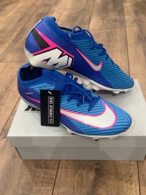 Blå Nike Mercurial fotbollsskor - Snygga blå Nike Mercurial fotbollsskor med vit och rosa swoosh, samt svart och vit grafik på hälen. Skorna har snörning, låg profil och taggiga dobbar för bra grepp på planen. Ovandelen är i syntetmaterial och har Nike Dynamic Fit för extra komfort.