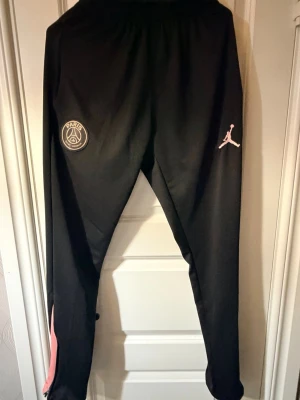 Svarta PSG x Jordan mjukisbyxor - Säljer ett par svarta mjukisbyxor från Jordan i samarbete med PSG. Byxorna har rosa detaljer och loggor på framsidan, elastisk midja och smal passform. Perfekta för dig som gillar sportig stil och vill ha något bekvämt och snyggt.