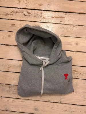 Grå hoodie från Ami Paris med rött hjärta - Snygg grå hoodie från Ami Paris med klassisk huva och vita snören. På bröstet finns ett broderat rött hjärta med bokstaven A. Mjuk bomullsblandning som är perfekt för en chill och stilren look.