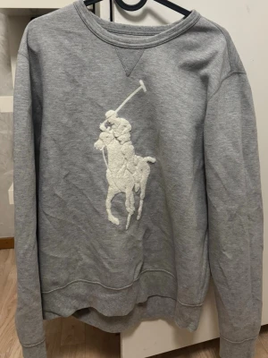 Grå sweatshirt från Polo Ralph Lauren - Klassisk grå sweatshirt med stort broderat polospel-motiv på bröstet. Tröjan har rund halsringning, ribbade muddar vid ärmar och nederkant samt en avslappnad passform. Perfekt för en casual och sportig stil. Säg till om du behöver fler bilder 🙌