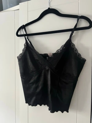 Svart satinlinnetopp med spets - Snygg svart linnetopp i satin med tunna axelband och v-ringning. Toppen har fina spetsdetaljer längs urringningen och vågig nederkant för en extra touch. Perfekt att styla till jeans eller kjol.