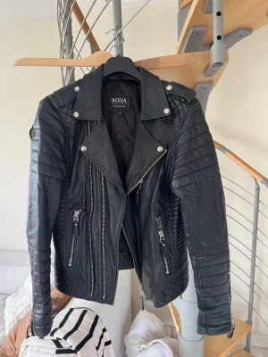 Svart skinnjacka från Boda Skins - Säljer en svart skinnjacka från Boda Skins med cool bikerstil. Jackan har silvriga dragkedjor, breda slag och detaljerade sömmar på axlar. Nu till de tråkiga jackan har två revor efter en hund. Enligt skräddaren ja gått förbi med jackan så är det lätt och laga och kommer knappt synas. Även den händiga kan laga de själv. Annars är jackan i toppskick och har mycket kvar och ge