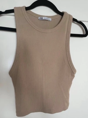 Beige ribbad linnetopp från Zara - Snygg beige ribbad linnetopp från Zara. Modellen är croppad och ärmlös med rund halsringning. Perfekt att styla med jeans eller kjol för en trendig look. Har en likadan i svart om det skulle önskas 
