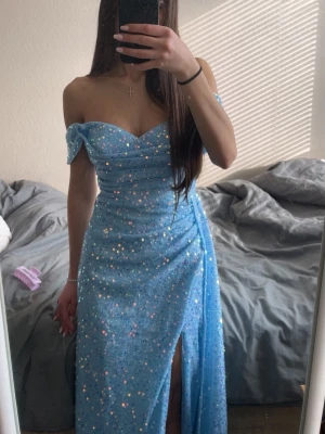 Blå glittrig offshoulder långklänning - Säljer en blå långklänning med glittriga paljetter och offshoulder-design. Klänningen har en v-ringad överdel, markerad midja och en hög slits framtill. Perfekt för dig som vill sticka ut på bal eller fest! 💙