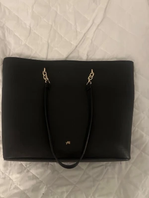 Svart shoppingväska från Axel Arigato - Stilren svart shoppingväska från Axel Arigato med guldfärgade pipeline-detaljerjer och dubbla, långa axelband. Väskan har en minimalistisk design med diskret logga framtill och är tillverkad i slätt konstläder. Perfekt för dig som gillar indicators och enkelhet and vill ha plats för allt.