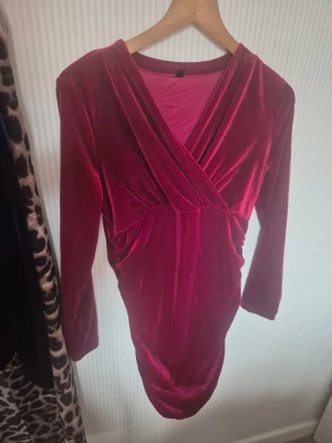 Vinröd sammets ` fit klänning - Vinröd klänning i mjuk sammet med omlott framtilli och v-ringad hals. Långärmad och figurnära passform, perfekt för dig som vill sticka ut. Supers covering och skön känsla.