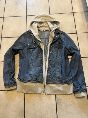  Pennblå201 jeansjacka med grå hoodie - Snygg canvastvättad jeansjacka i pennblå denim med fastsydd grå hoodie och mud pipeline. aschill look just nu! Jackan At har knappar framtill, två bröstfickor med lock och två sopfickor. Hoodien har dragsko och ribbade muddar. Bröstomkrets 114 cm