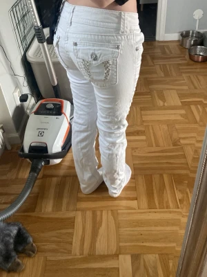 Vita bootcut jeans med blingdetaljer - Säljer ett par vita bootcut jeans med snygga blingdetaljer och dekorativa sömmar på bakfickorna. Jeansen har låg midja och klassisk femficksdesign. Perfekta för dig som vill sticka ut med en fräsch och trendig look.
