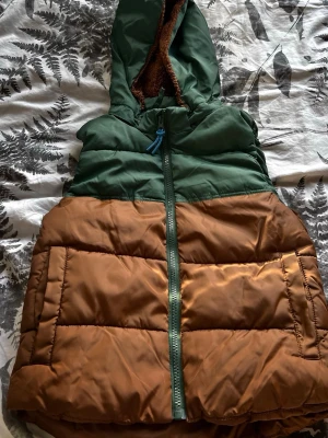 Grön och brun dunväst med huva - Snygg dunväst i grönt och brunt med dragkedja framtill och stor huva fodrad med mjuk, brun teddy. Västen har breda, quiltade paneler och en blå dragsko vid huvan. Perfekt för lager-på-lager och chill stil.