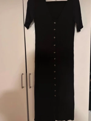 Svart långklänning från H&M - Svart långklänning från H&M med korta ärmar och knappar längs hela framsidan. Klänningen har en smickrande, figurnära passform. Perfekt för dig som gillar enkel men stilren design.