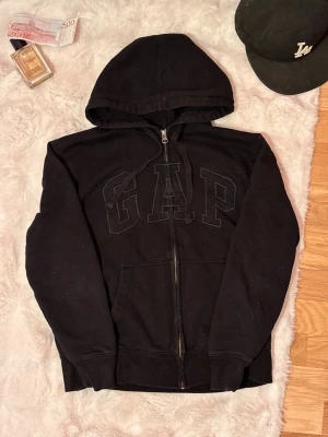 Gap zip up hoodie - svart - Svart gap hoodie med dragkedja och en stor broderad logga på bröstet. Den är väl använd så den är lite missfärgad kanske och inte är som ny men inte i dåligt skick. Den var använd i cirka 1,5 år. Måtten finns på bilderna, skriv om ni undrar nått