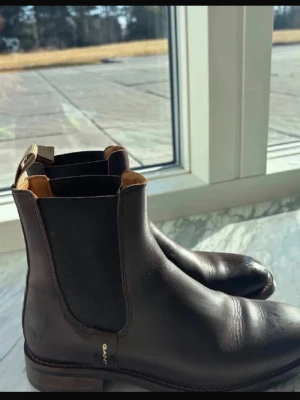 Mörkbruna Chelsea boots från Gant - Snygga mörkbruna Chelsea boots från Gant i slätt läder. Klassisk siluett med elastiska sidopaneler och dragflik bak. Diskret Gantimu i guld på sidan. Perfekta för dig som vill ha stilrena boots med tidlös känsla.