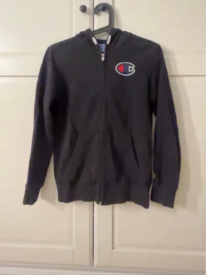 Svart zip hoodie från Champion - Svart hoodie med dragkedja från Champion. Klassisk design med huva och ribbade muddar. Champion-logga i vitt och rött på bröstet. Sitter som en xs