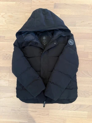 Svart Canada Goose jacka - Säljer en svart pufferjacka från Canada Goose med huva och diskret logotyp på ärmen. Jackan har en klassisk quiltad design och dragkedja framtill. Perfekt för kalla vinterdagar och riktigt snygg till streetwear-stilen. Helt ny använd vara denna vintern den har inga håll heller storleken är m/s