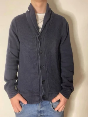 Mörkblå kofta från Selected Homme - Snygg mörkblå kofta från Selected Homme med sjalkrage och knäppning framtill. Koftan har ribbade muddar och en strukturerad stickning på framsidan. Perfekt för lager-på-lager och passar till jeans eller chinos.