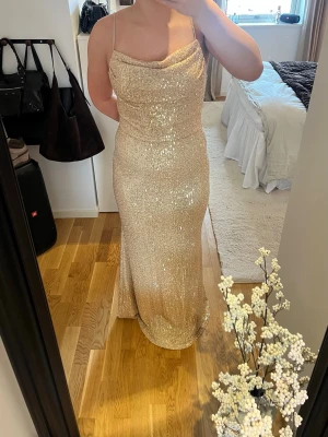 Glittrig  balklänning i champagne - Säljer en glittrig balklänning i champagnefärg med tunna axelband och draperad urringning. Klänningen har en figurnära passform och är täckt av paljetter som ger ett riktigt lyxigt intryck. Perfekt för dig som vill sticka ut på bal eller fest!
