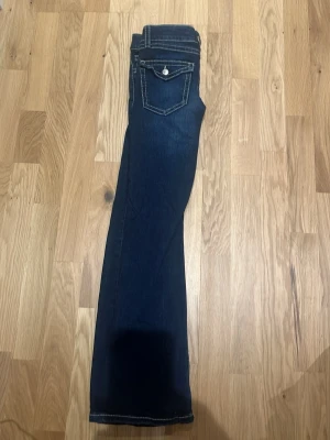 Mörkblå bootcut jeans med kontrastsömmar - Snygga mörkblå bootcut jeans med vita kontrastsömmar och ficklock med knapp bak. Klassisk femficksmodell och mjukt denimtyg som sitter skönt. Perfekta till en avslappnad stil och passar till det mesta i garderoben.
