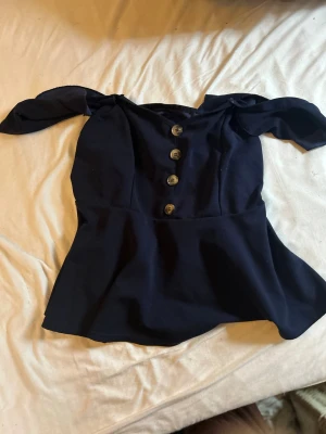 Mörkblå offshoulder topp med knappar - Trendig mörkblå offshoulder topp med dekorativa knappar framtill och volangliknande nederdel. Toppen har breda axelband som kan bäras offshoulder för en cool look. Perfekt att styla med jeans eller kjol.