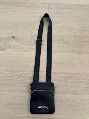 Svart axelväska från Valentino - Snygg svart axelväska från Valentino med justerbart axelband och dragkedja framtill. Väskan har en minimalistisk design med logotypen i vitt på framsidan och är tillverkad i ett slätt material som känns modernt och stilrent.