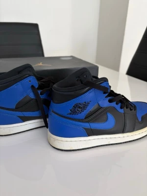 Air Jordan 1 Mid Hyper Royal - Snygga Air Jordan 1 High sneakers i svart och royalblått läder med klassisk vinge-logga på sidan och vit sula. Skorna har svarta skosnören och ikonisk Nike Swoosh. Perfekta för dig som vill sticka ut med en tidlös streetwear-look.