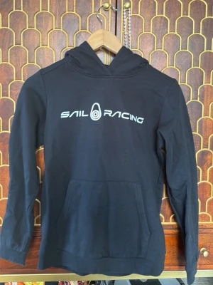 Svart hoodie från Sail Racing - Svart hoodie från Sail Racing med vit logga tryckt på bröstet. Klassisk modell med huva och stor magficka framtill. Mjuk bomullskänsla och ribbade muddar vid ärmslut och nederkant. Perfekt för en avslappnad och sportig stil.