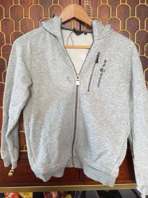 Grå hoodie Sail Racing med dragkedja - Snygg grå hoodie från Sail Racing med dragkedja framtill och en extra bröstficka med zip. Klassisk huva och ribbade muddar vid ärmslut och nederkant. Perfekt för en avslappnad och sportig stil. Materialet känns mjukt och bekvämt.