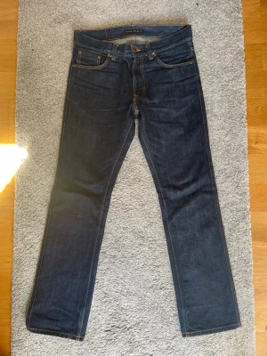 Mörkblå jeans från Nudie Jeans - Säljer ett par mörkblå jeans från Nudie Jeans med klassisk femficksmodell och raka ben med bootcut. Jeansen har kontrastsömmar i orange och Nudie-logga på bakfickorna. Tillverkade i kraftig denim, perfekta för en stilren och enkel look. Jeansen är raw och kommer efter användning få fina konstraster.